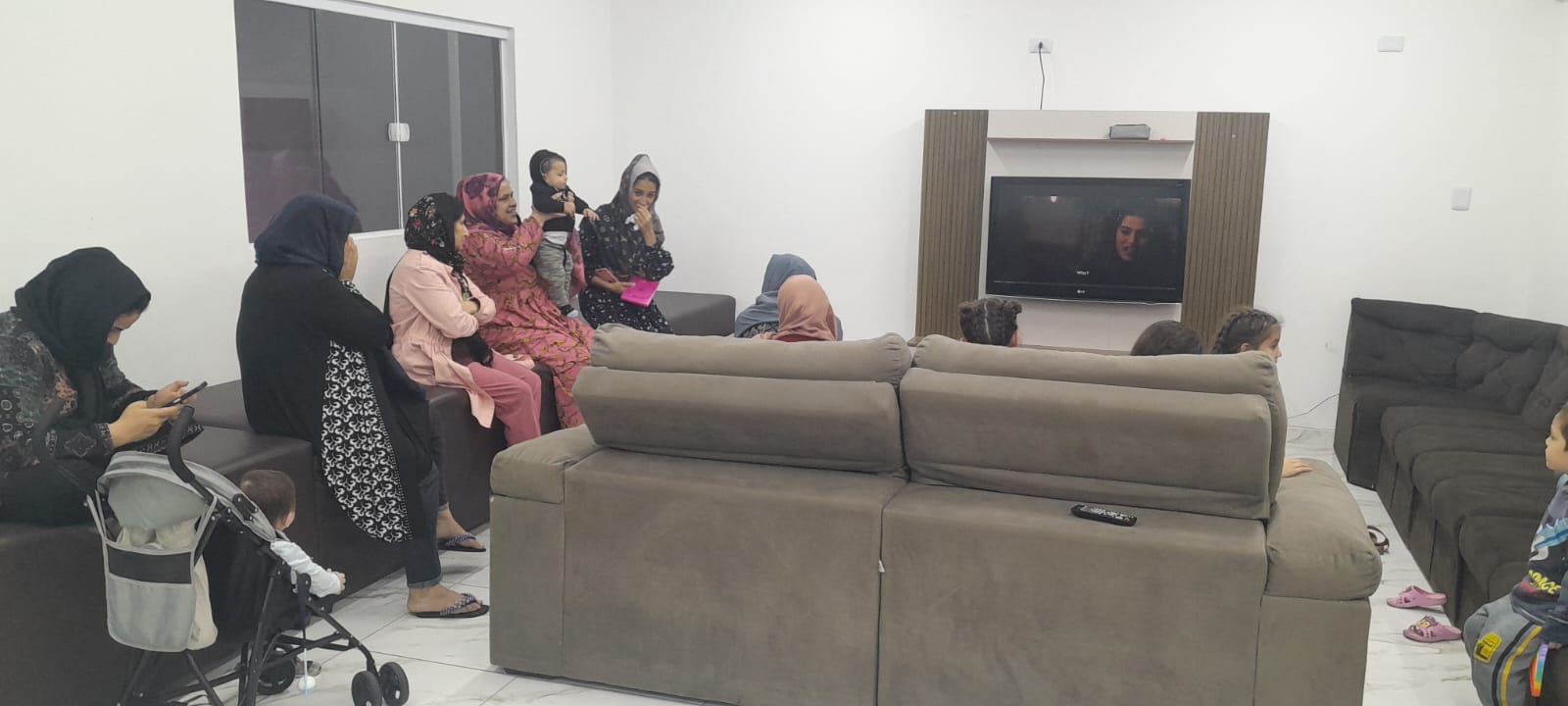 Na foto, vemos mulheres e seus filhos sentadas ao redor de uma televisão, um sofá marrom de costas para a imagem, com cinco mulheres sentadas a esquerda da imagem. A primeira, veste roupas escuras e possui um carrinho de bebê ao lado, a direita dela, outra de vestimentas escuras, seguida por uma de rosa claro, uma de rosa escuro segurando uma criança e outra de roupas escuras e touca. No centro da sala, uma televisão posicionada em um móvel de madeira na parede.